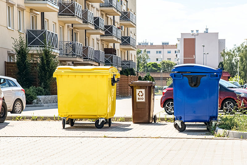 Ein gelber und ein blauer Müllcontainer. Dazwischen steht eine braune Mülltonne.